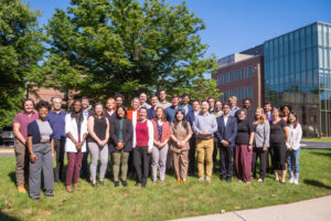 New Faculty pose for a group photo in front of a tree and new building.